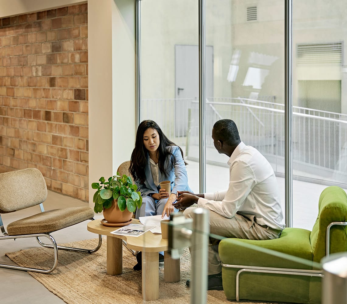 Two people sit on green chairs in a modern office space, discussing documents on a small wooden table. One holds a cup, and a potted plant is on the table. Large windows provide natural light.